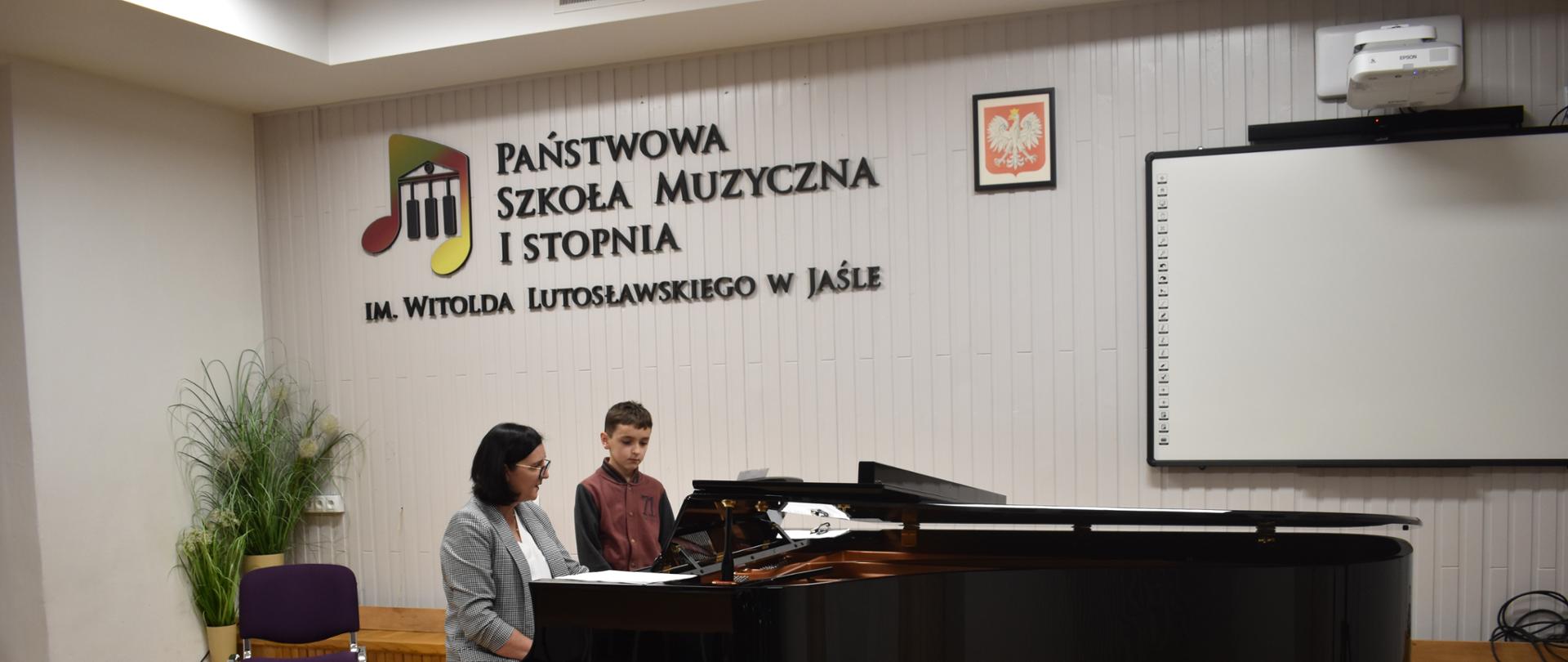 Na zdjęciu widnieje nauczyciel fortepianu mgr Dorota Skibicka, która w auli szkoły instruuje ucznia jak grać na fortepianie. Tło zdjęcia jest białe i jasno brązowe.