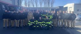 Grupowe zdjęcie 29 strażaków z województwa opolskiego biorących udział w szkoleniu dotyczącym działań ratowniczych przy pojazdach elektrycznych i hybrydowych, prowadzonym przez instruktorów z Republiki Czeskiej. Uczestnicy stoją przed budynkiem strażnicy i samochodami pożarniczymi, ubrani w mundury służbowe oraz odzież ochronną.
