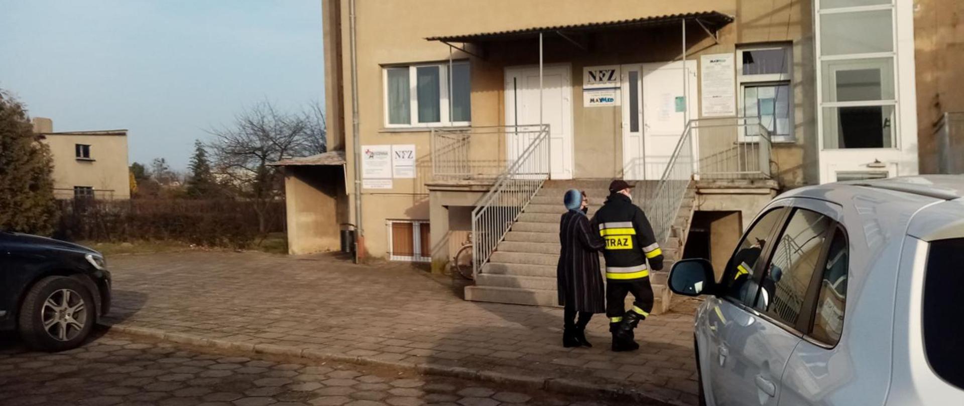 Strażacy pomagający starszej kobiecie w wejściu po schodach do punktu szczepień