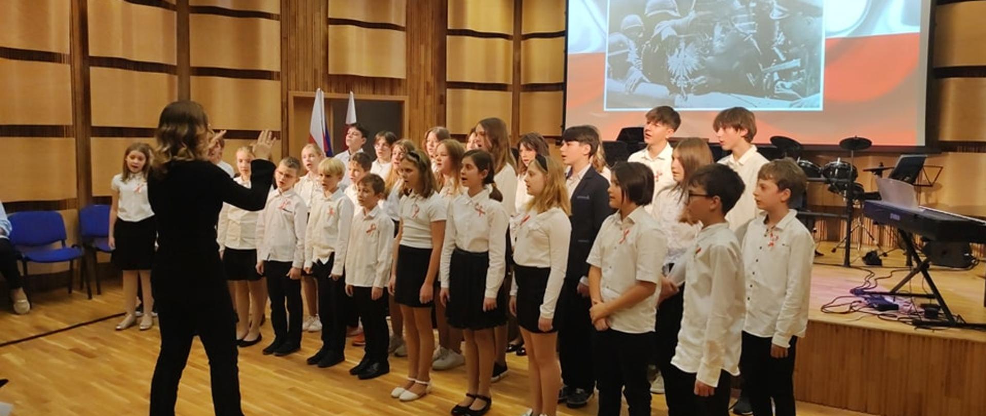 Zdjęcie chóru dzieci z dyrygentem w sali koncertowej. W tle na ekranie flaga z żółnierzami. Koncert z Okazji Narodowego Dnia Niepodległości.