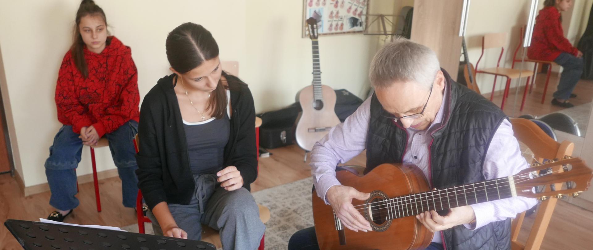 Nauczyciel prowadzący warsztaty gra na gitarze uczennicy. Dziewczyna obserwuje grę. Za nią siedzi dziewczyna obserwująca lekcję.