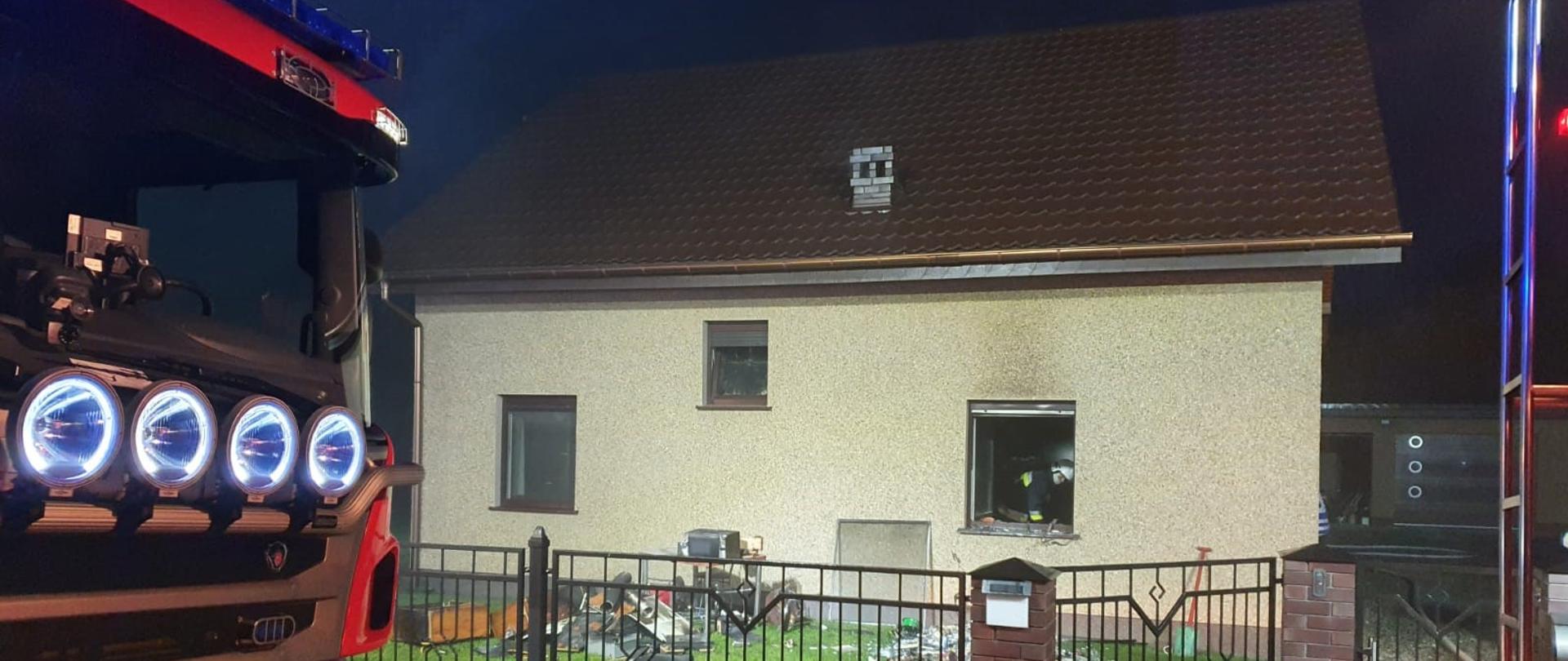 Na zdjęciu na drugim planie widać budynek mieszkalnym, w którym doszło do pożaru. Obiekt w kolorze beżowym. Dach koloru brązowego. W prawej części budynku widać otwarte okno, wewnątrz strażak w ubraniu specjalnym, który wyrzuca przez okno nadpalone elementy wyposażenia. Zdjęcie wykonane z ulicy w porze nocnej. Na pierwszym planie z lewej strony widać przód samochodu gaśniczego z włączonymi światłami. Po prawej stronie tył drugiego samochodu gaśniczego. Między samochodami a obiektem mieszkalnym znajduje się płot w kolorze czarnym i brązowym oraz zielona trawa.