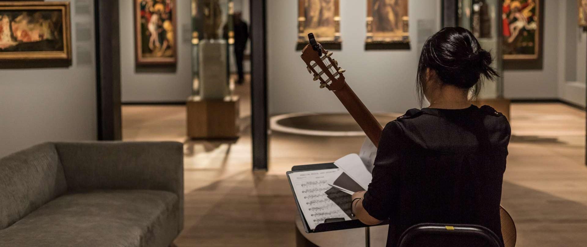 zdjęcie przedstawiające pomieszczenie galerii z obrazami zawieszonymi na szarych ścianach oraz ciemnowłosą kobietę odwróconą tyłem, która siedzi na krześle z gitarą w ręku, po lewej stronie znajduje się pulpit z nutami leżącymi na pulpicie 