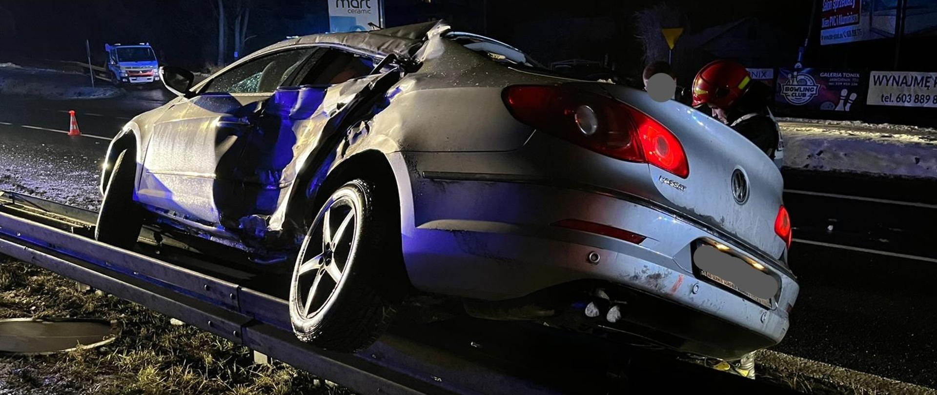 Zdjęcie wykonane w porze nocnej przedstawia uszkodzony samochód osobowy, który w wyniku zderzenia najechał i zatrzymał się na barierach ochronnych rozdzielających pasy ruchu drogi krajowej. Częściowo widoczny strażak wykonuje czynności opisujące zdarzenie. W tle widoczne banery reklamowe i stojące auto pomocy drogowej.