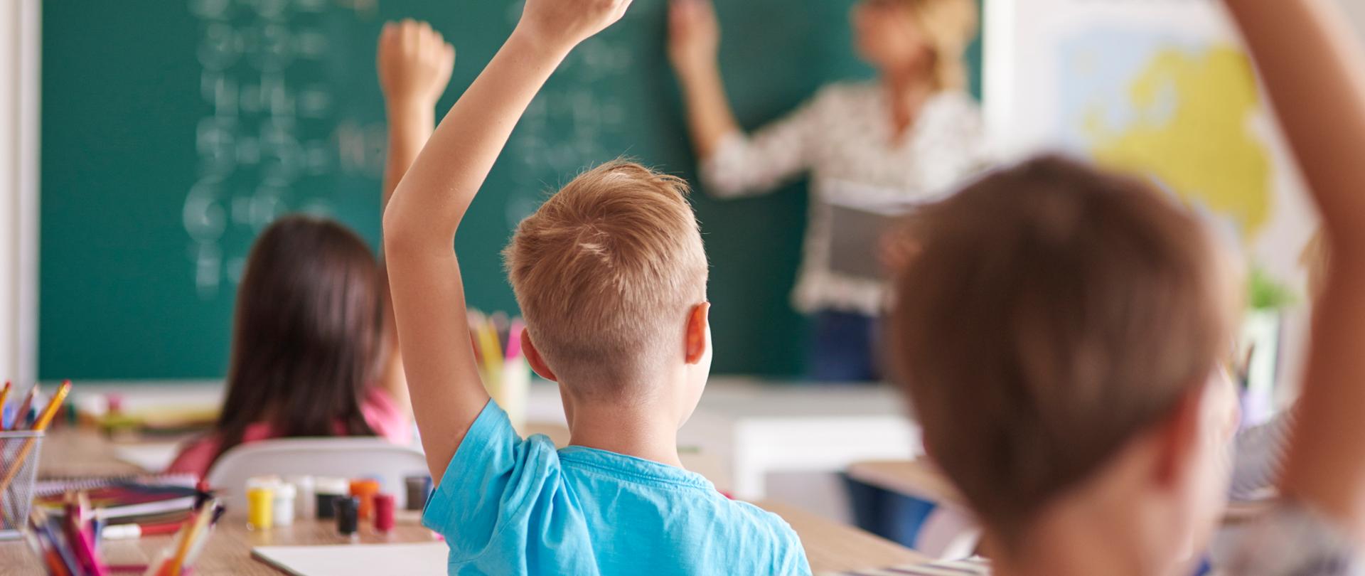 W klasie szkolnej kilkoro dzieci siedzi w ławkach i zgłasza się do odpowiedzi, unosząc ręce do góry. Na pierwszym planie widać chłopca w niebieskiej koszulce. W tle stoi nauczycielka, która pisze na zielonej tablicy. 