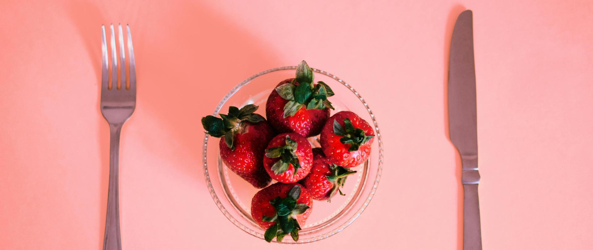 Czerwone truskawki z zielonymi szypułkami w szklanej przezroczystej salaterce. Po lewej stronie miseczki nóż, po prawej widelec.