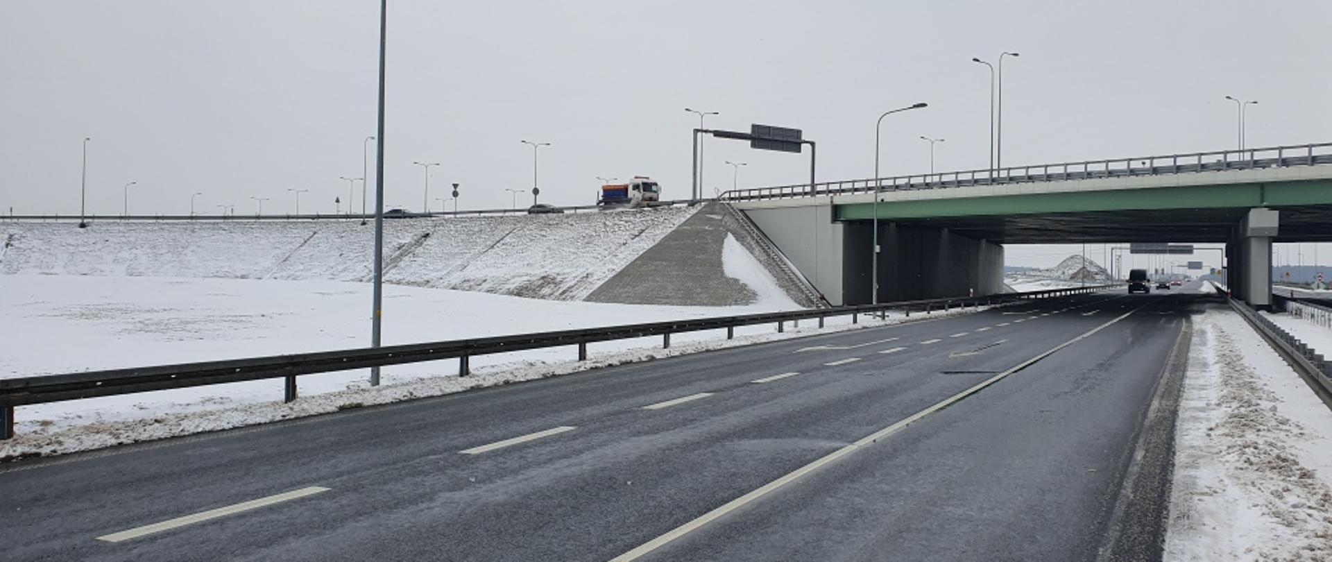 Zdjęcie przedstawia drogę ekspresową zimą. Nawierzchnia drogi czarna, mokra. W tle wiadukt drogowy, po którym porusza się pojazd specjalistyczny do zimowego utrzymania dróg - pługosolarka. W tle widoczne również ośnieżone pobocza i pola.