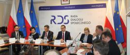 Na zdjęciu widnieje stół prezydialny za którym siedzi od lewej strony: Waldemar Buda - Minister Rozwoju i Technologii, Marlena Maląg - Minister Rodziny i Polityki Społecznej, Łukasz Bernatowicz - Przewodniczący rady Dialogu Społecznego, Premier Mateusz Morawiecki - Prezes Rady Ministrów, Anna Moskwa - Minister Klimatu i Środowiska, Rafał baniak - Prezes Zarządu Pracodawców RP. W tle siedzących baner z napisem Rada Dialogu Społecznego. Po lewej stronie trzy flagi: UE, Polski i UE. oraz po prawej stronie też takie sam układ flag. nad banerem wisi godło Polski. 