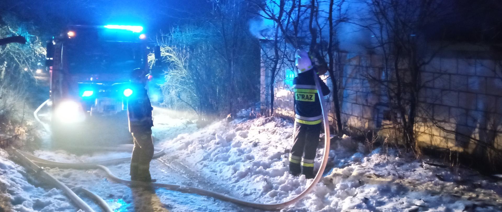 Zdjęcie zrobione w nocy. Na pierwszym planie dwóch strażaków w ubraniach specjalnych. Jeden trzyma węża i gasi pożar. Drugi asekuruje go. W tle stoi samochód strażacki na sygnałach błyskowych. Z budynku po prawej wydobywa się dym.