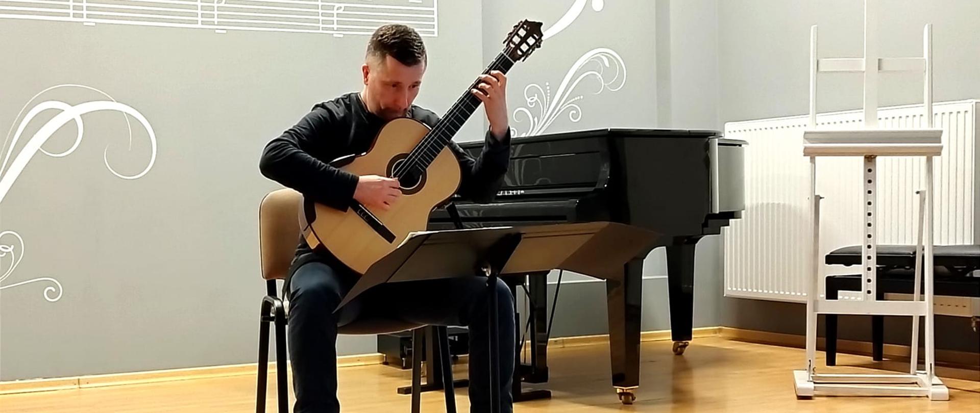 Na zdjeciu Waldemar Gromolak podczas koncertu trzymający gitarę. W tle stoi fortepian