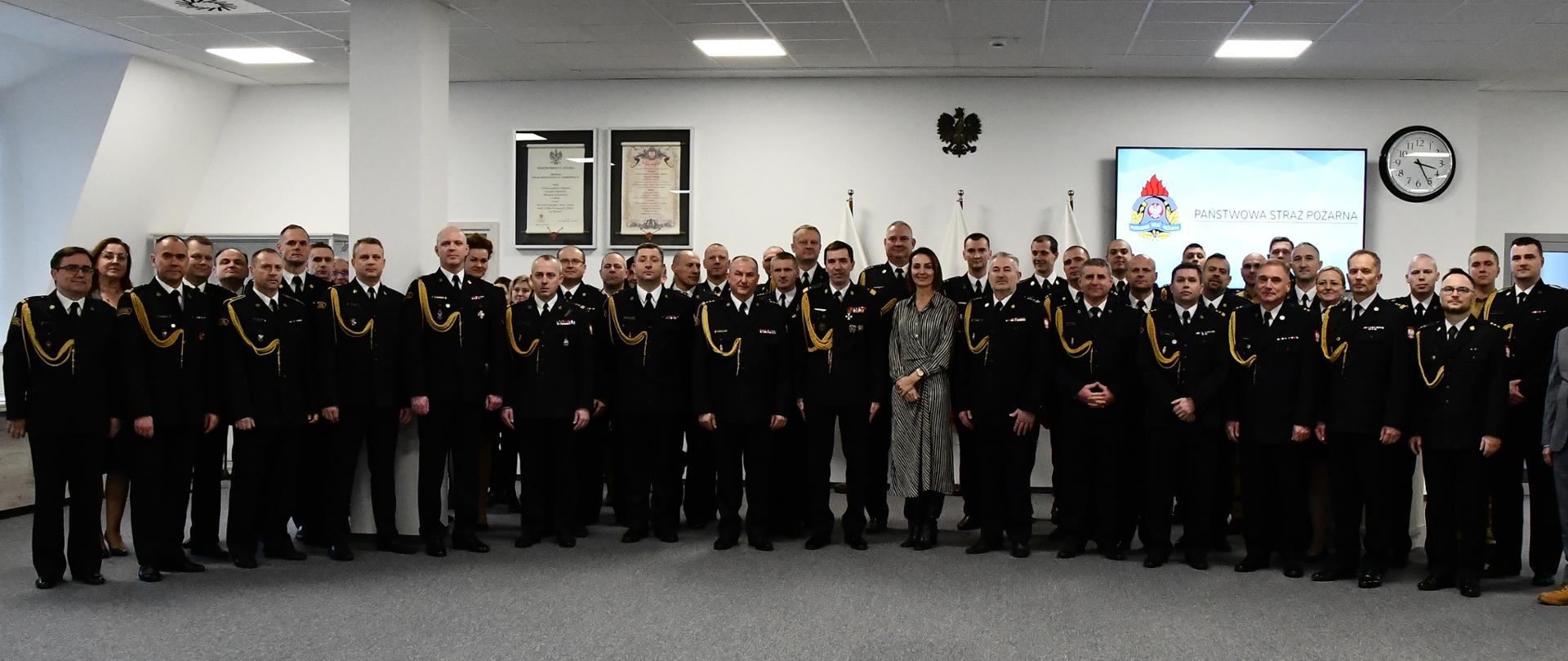 Strażacy w mundurach wyjściowych pracownicy cywilni wojewoda pomorska stoją obok siebie za nimi na ścianie wiszą rota ślubowania ustawione są flagi Polski godło ekran z logiem i napisem Państwowa Straż Pożarna zegar krzyż oraz znak wyjścia ewakuacyjnego. 