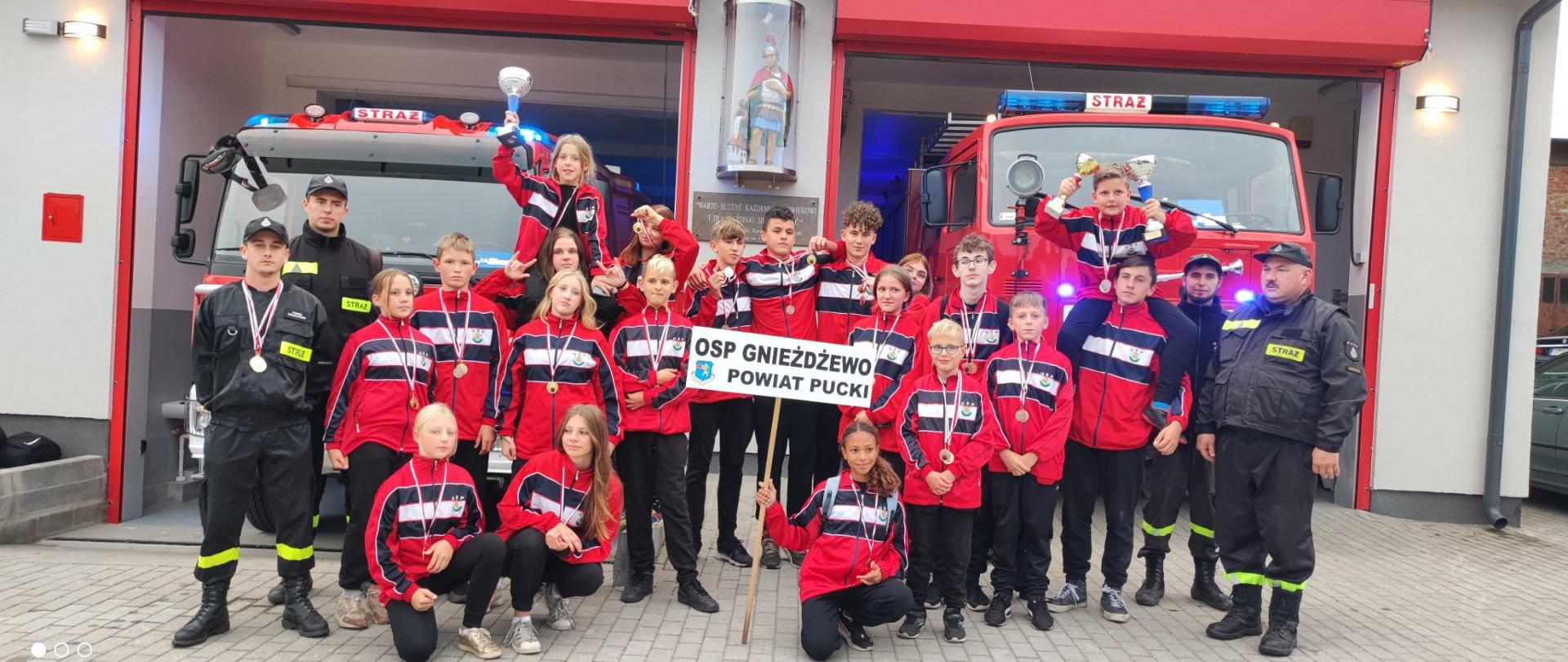 Na zdjęciu widoczne są dzieci z młodzieżowej drużyny pożarniczej. Ubrani w czerwono czarne dresy, trzymają puchary i medale. W tle samochody pożarnicze.