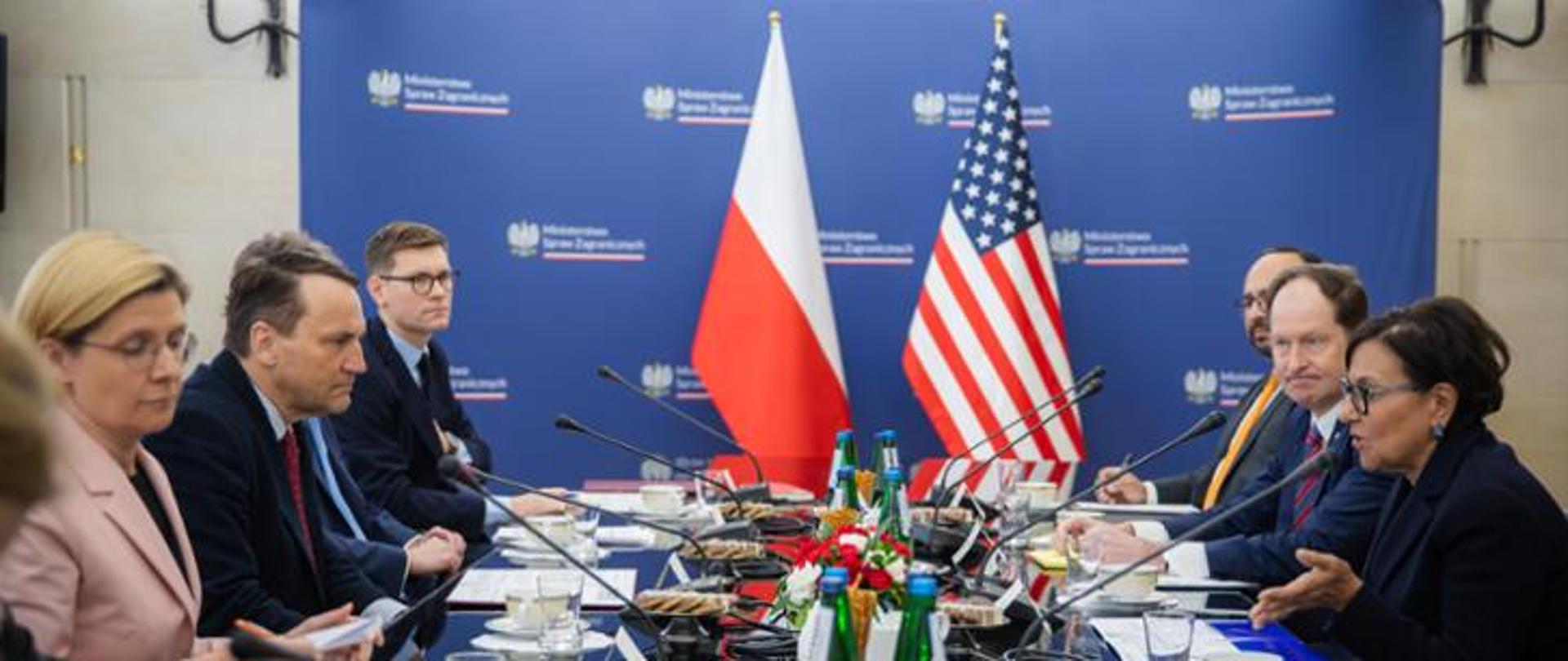 People are sitting at the table. Flags of the USA and Poland are in the background. 