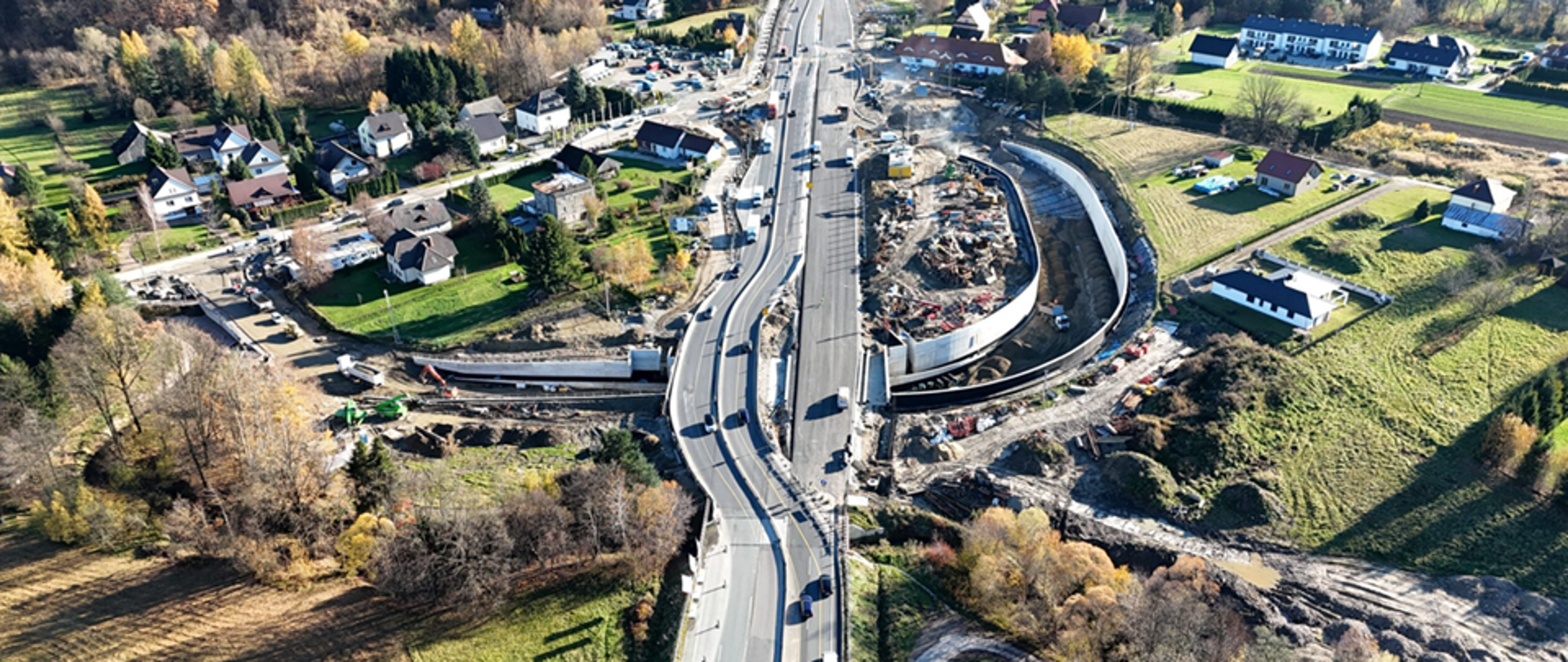 Zdjęcie przedstawia widok z lotu ptaka na rozległy teren wiejski z przebiegającą przez środek główną drogą. Po obu stronach drogi znajdują się domy jednorodzinne z ogrodami oraz pola uprawne. W centralnej części obrazu trwa duża budowa – widoczne są wykopy, maszyny budowlane i betonowe konstrukcje. Teren jest otoczony zielenią, drzewami w jesiennych barwach oraz fragmentami lasu w tle.