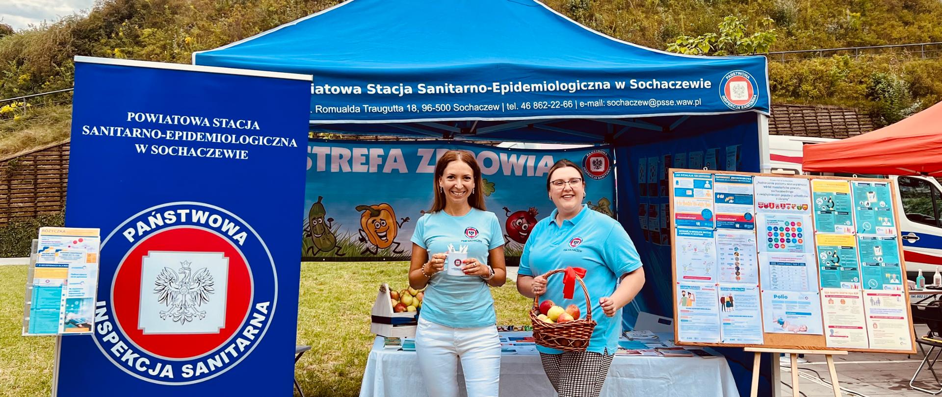 Dwie pracownice PSSE w Sochaczewie w błękitnych koszulkach z logiem Inspekcji Sanitarnej przed stoiskiem prozdrowotnym podczas 8. Święta Bzury w Sochaczewie. Jedna z kobiet trzyma w rękach koszyk z jabłkami, druga kubek z długopisami promocyjnymi. W tle widać Ruiny Zamku Książąt Mazowieckich w Sochaczewie.