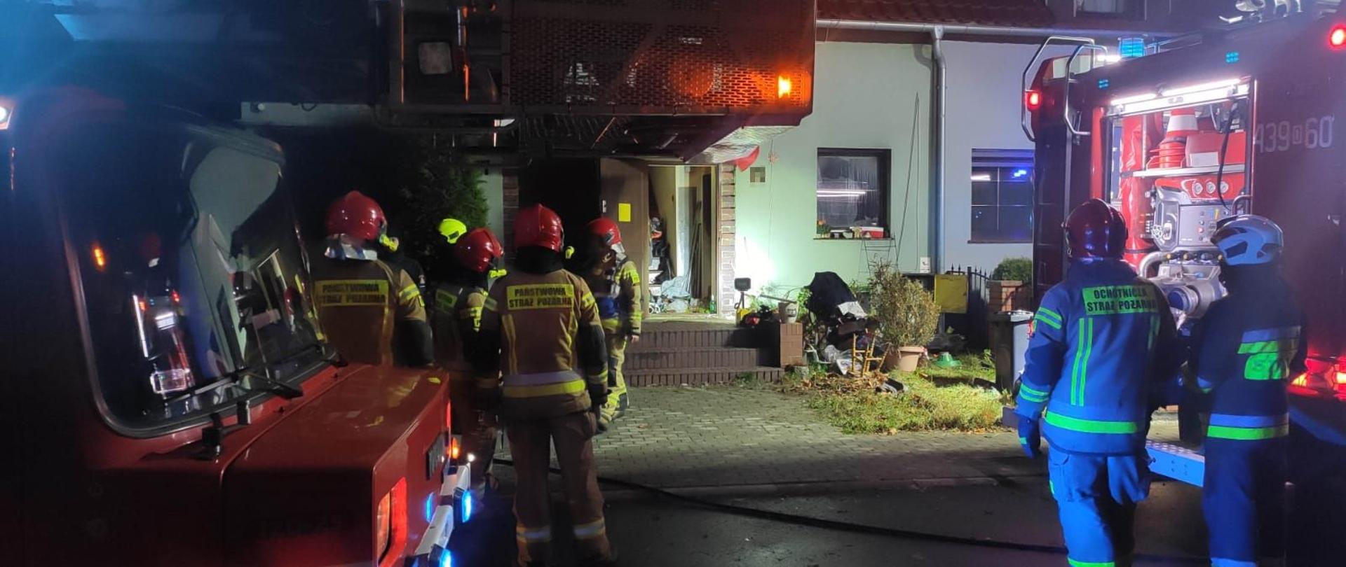 Strażacy Państwowej Straży Pożarnej oraz OSP w ubraniach specjalnych stojący przy samochodach pożarniczych przed budynkiem mieszkalnym, w którym doszło do pożaru mieszkania. W budynku otwarte drzwi wejściowe na oścież.