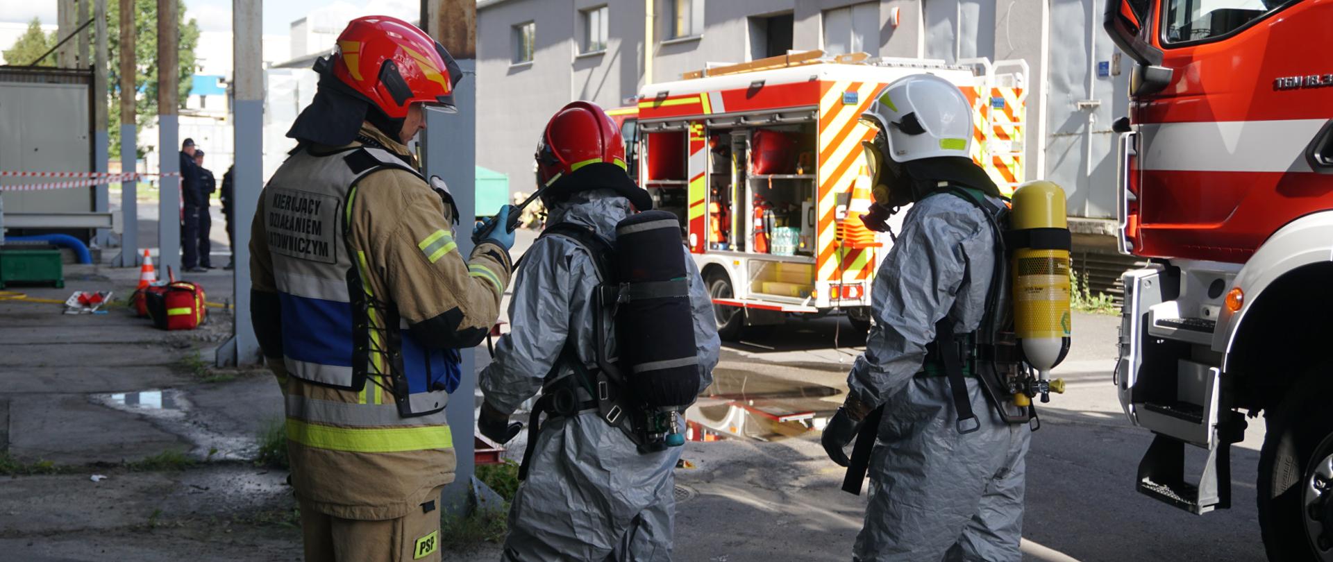 W centralnej części znajduje się dwóch strażaków ubranych w specjalne kombinezony chroniące przed skarżeniem chemicznym w kolorze szarym. Mają oni na sobie aparaty ochrony dróg oddechowych oraz hełmy. Tuż obok po lewej stronie znajduję się dowódca ubrany w niebiesko-szarą kamizelką „ kierujący działaniem ratowniczym”, trzyma on w ręce radiotelefon. W oddali widać samochód strażacki, który ma pootwierane skrytki. W lewej części kadru widać miejsce do którego zmierzają strażacy, w oddali widać torbę medyczną i nosze.