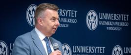 Minister Wieczorek stoi na tle granatowej ścianki z napisem Uniwersytet Kazimierza Wielkiego w Bydgoszczy i mówi do trzymanego w ręku mikrofonu.