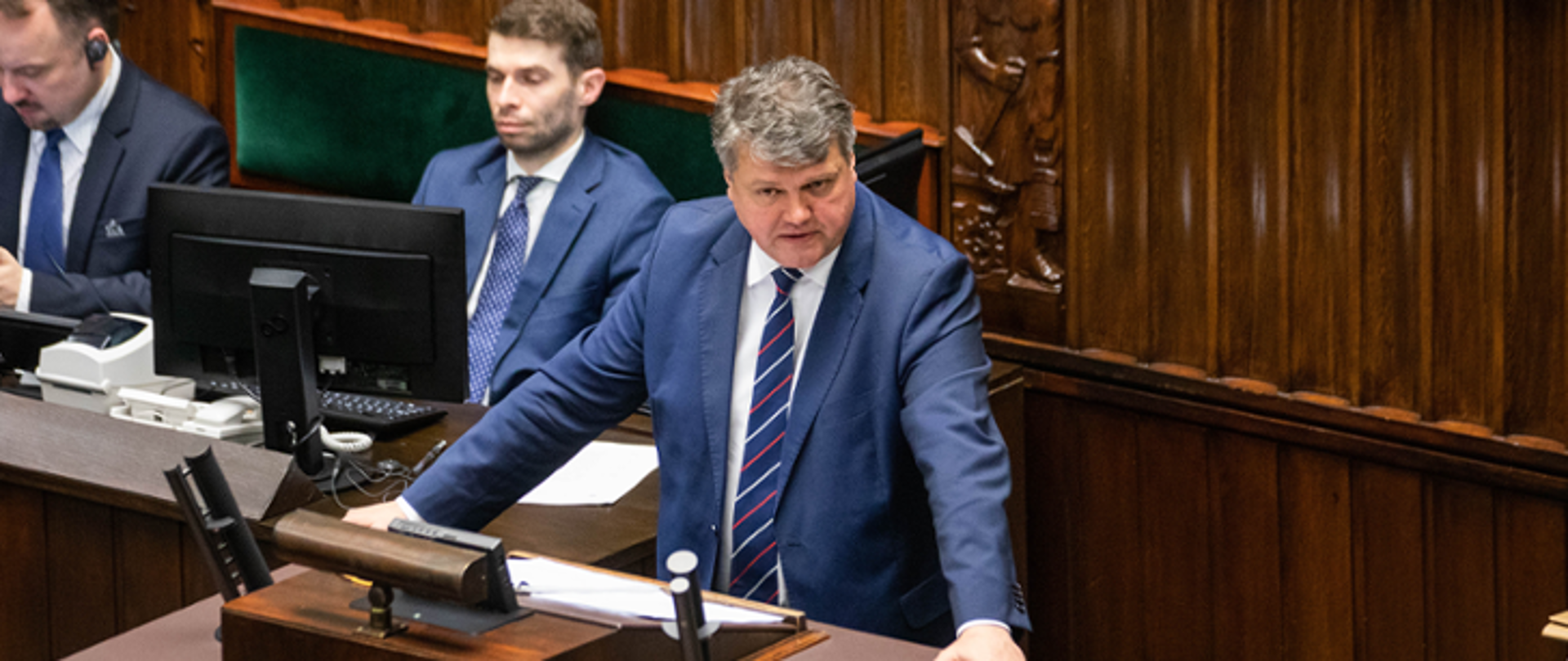 Jeden mężczyzna stojący przy mównicy i wypowiadający się. Za nim siedzi dwóch mężczyzn