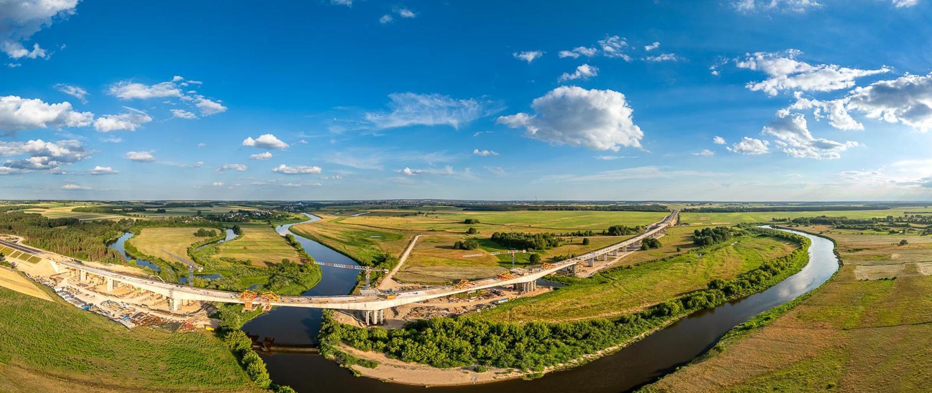 Widok z lotu ptaka na wielką plac budowy mostu nad meandrującą rzeką. Rzeka wygina się przez krajobraz, zielone pola i skupiska drzew po obu stronach. Na placu budowy widać wiele żurawi, kilka częściowo ukończonych odcinków mostu z widocznymi rusztowaniami oraz liczne pojazdy budowlane. Niebo jest bezchmurne, co sugeruje dobre warunki pogodowe do prac budowlanych.