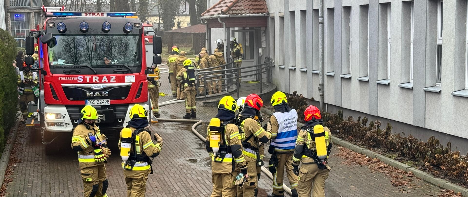 Strażacy prowadzą działania przed budynkiem. Na zdjęciu są strażacy oraz pojazd pożarniczy.
