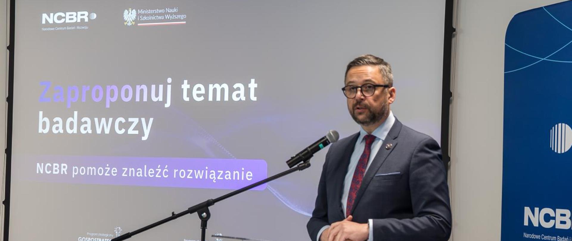 Zdjęcie pod kątem z boku, na tle ekranu z napisem Zaproponuj temat badawczy - NCBR pomoże znaleźć rozwiązanie za mównicą stoi minister Kulasek i mówi do mikrofonu na stojaku.