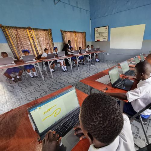 W sali lekcyjnej uczniowie i uczennice siedzą naprzeciwko siebie w dwóch rzędach i pracują na komputerach