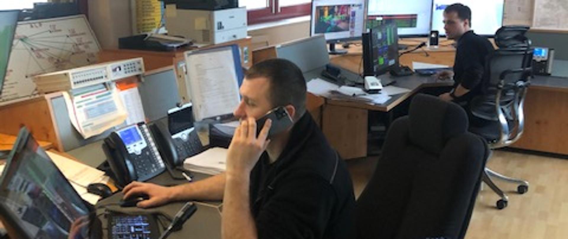 Zdjęcie przedstawia pomieszczenie Stanowiska Kierowania Komendanta Powiatowego Państwowej Straży Pożarnej w Międzyrzeczu. Dwóch dyżurujących funkcjonariuszy prowadzi obsługę zgłoszeń związanych z silnymi wiatrami.