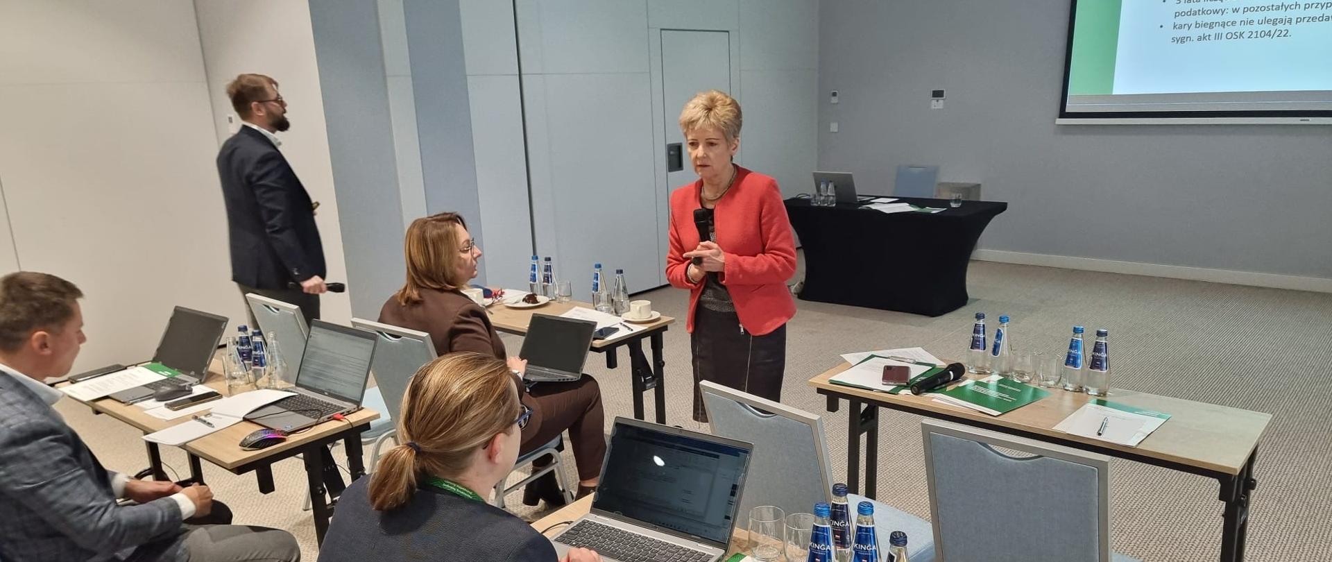 Minister Hanna Kończal, Główny Inspektor Ochrony Środowiska, prowadzi naradę. Kilkoro uczestników narady siedzi przy stołach z laptopami i notatkami. Na ekranie wyświetlony jest slajd z treścią dotyczącą postępowań prawnych.