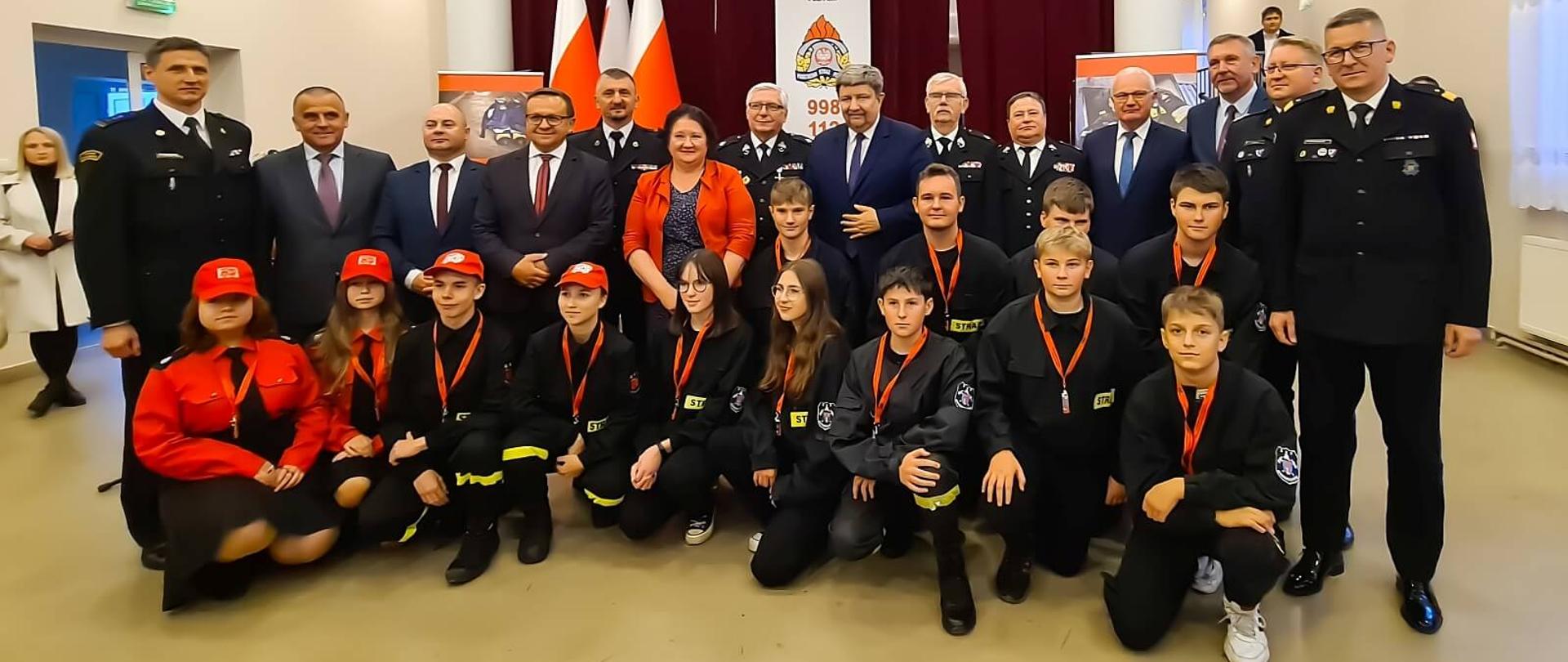 uczestnicy uroczystości wręczenia promes dla młodzieżowych drużyn pożarniczych pozują do zdjęcia zbiorowego wśród nich dzieci z młodzieżowej drużyny pożarniczej, strażacy druhowie, posłowie i senatorowie RP, przedstawiciele władz samorządowych
