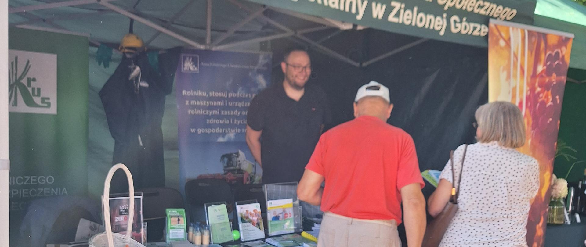 Przy zielonym namiocie stoją kobieta i mężczyzna. Pod namiotem stoisko z materiałami informacyjnymi KRUS, za stołem stoi mężczyzna – pracownik KRUS.