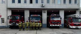 Zdjęcie przedstawia strażaków na tle samochodów oraz garaży jednostki ratowniczo-gaśniczej.