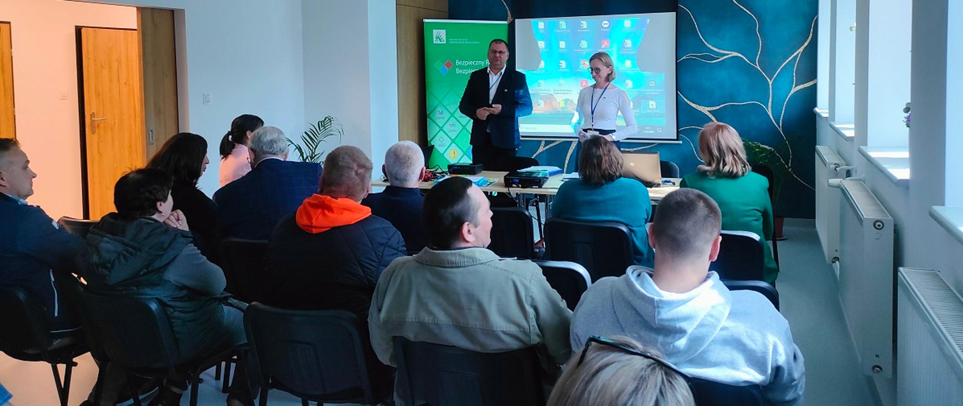 W sali konferencyjnej urzędu na krzesłach ustawionych w rzędach, siedzi grupa uczestników, zwróconych w stronę prowadzących. Przodem do nich stoją dwie osoby – mężczyzna i kobieta. Za nimi znajduje się ekran projekcyjny z prezentacją. Za mężczyzną stoi zielony roll-up z widocznym logiem KRUS.