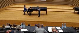 Chłopiec stojąc na środku estrady sali koncertowej PSM gra na flecie poprzecznym, za nim na fortepianie gra mężczyzna. Z przodu widać od tyłu widownię oraz siedzące przy stole Jury.