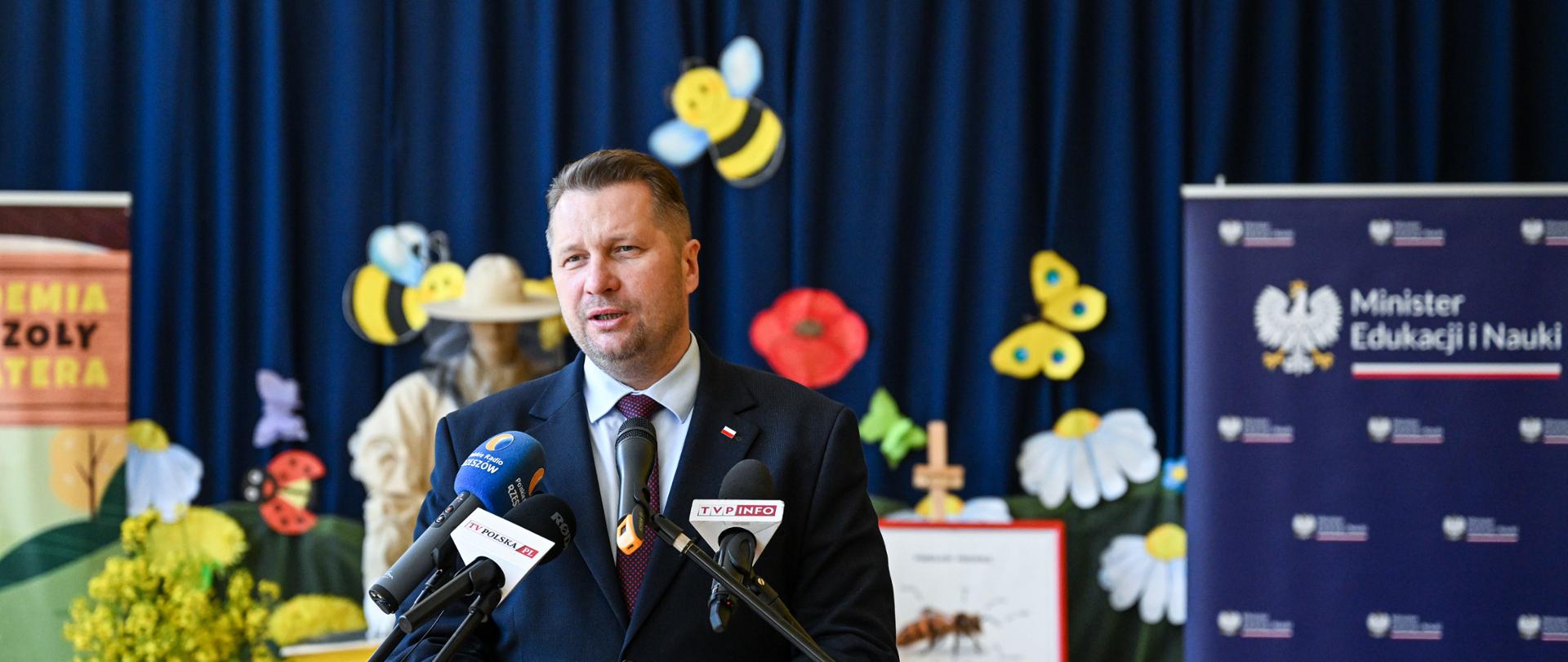 Minister Czarnek stoi za mównicą, mówi do kilku mikrofonów na stojakach, za nim ściana zasłonięta granatowym materiałem z żółtym napisem "Akademia pszczoły bohatera".