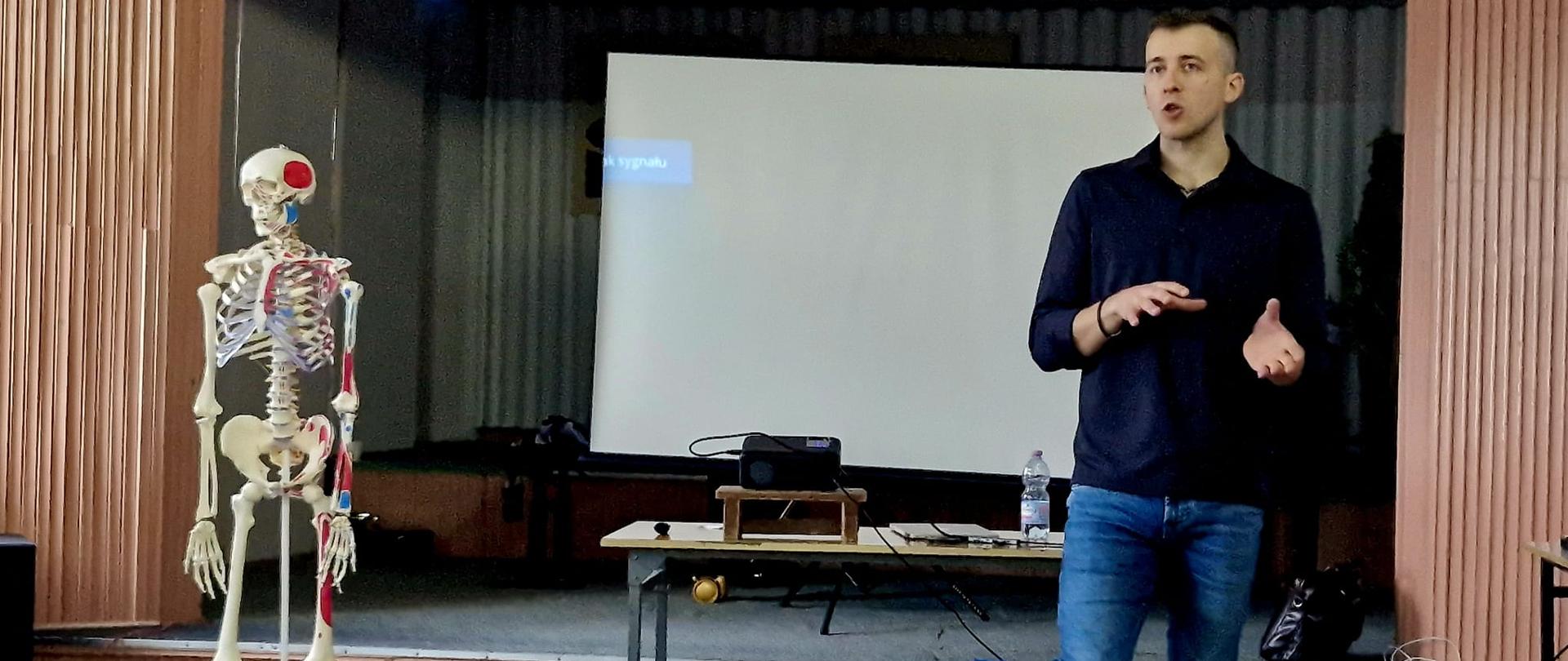 Mężczyzna - fizjoterapeuta prowadzący wykład. Z lewej strony pomoc dydaktyczna - fantom szkieletu