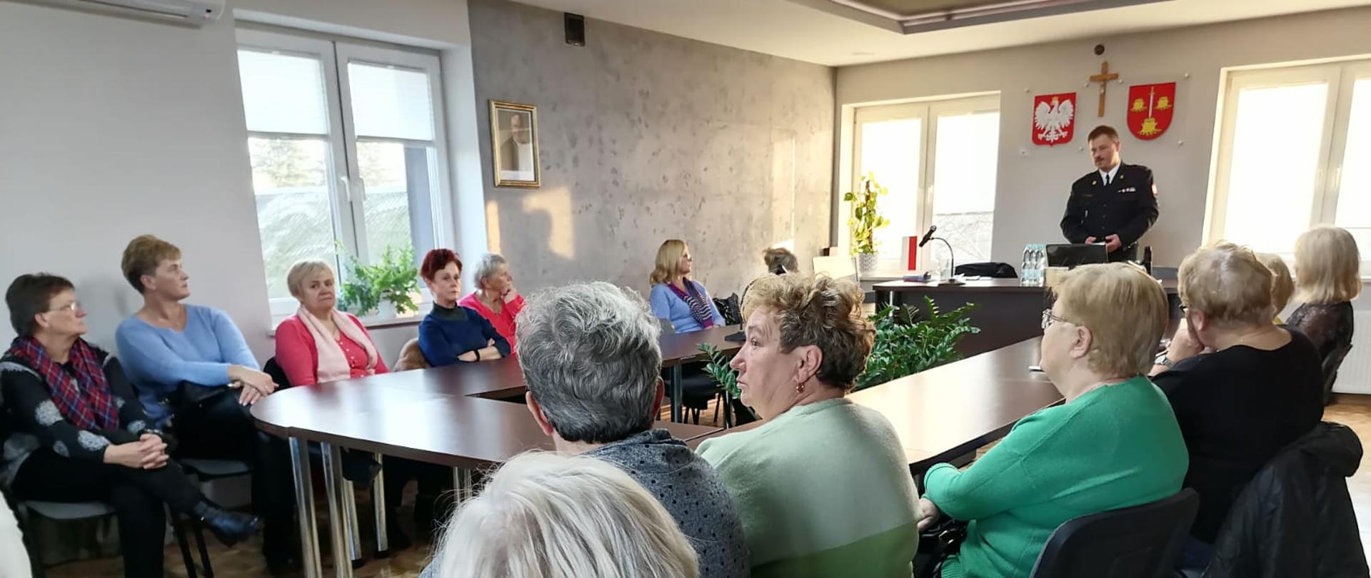 Zdjęcie wykonane wewnątrz sali konferencyjnej urzędu gminy w Goździe. Na zdjęciu widać stół. Dookoła niego siedzą kobiety. Po prawej stronie stołu stoi funkcjonariusz Państwowej Straży Pożarnej. Strażak ubrany jest w mundur galowy w ciemnym kolorze. Funkcjonariusz wygłasza pogadankę w ramach akcji w ramach wspólnych działań prewencyjnych o zasadach zachowania bezpieczeństwa na drodze pieszych i rowerzystów w ramach akcji "Świeć przykładem - i Ty kiedyś będziesz seniorem" a także o zagrożeniach związanych z występowaniem tlenku węgla podczas użytkowania urządzeń grzewczych w ramach akcji "Czujka na straży Twojego bezpieczeństwa".
