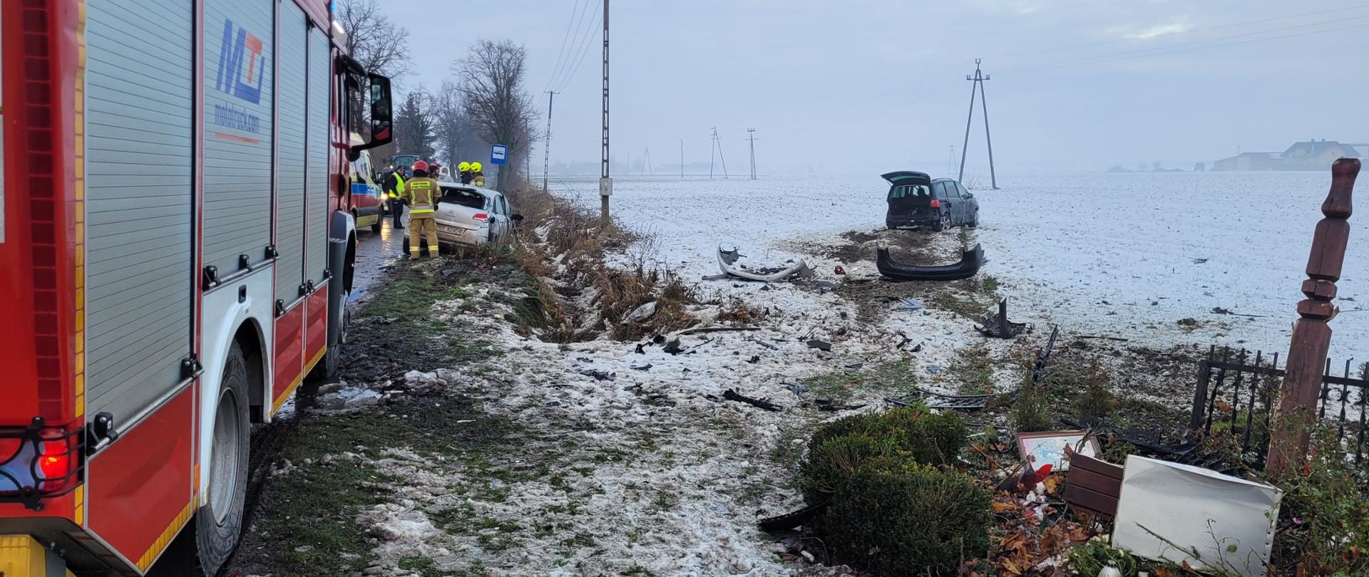 Zdjęcie przedstawia zdeformowane bryły pojazdów na polu pokrytym warstwą śniegu na tle pojazdu pożarniczego