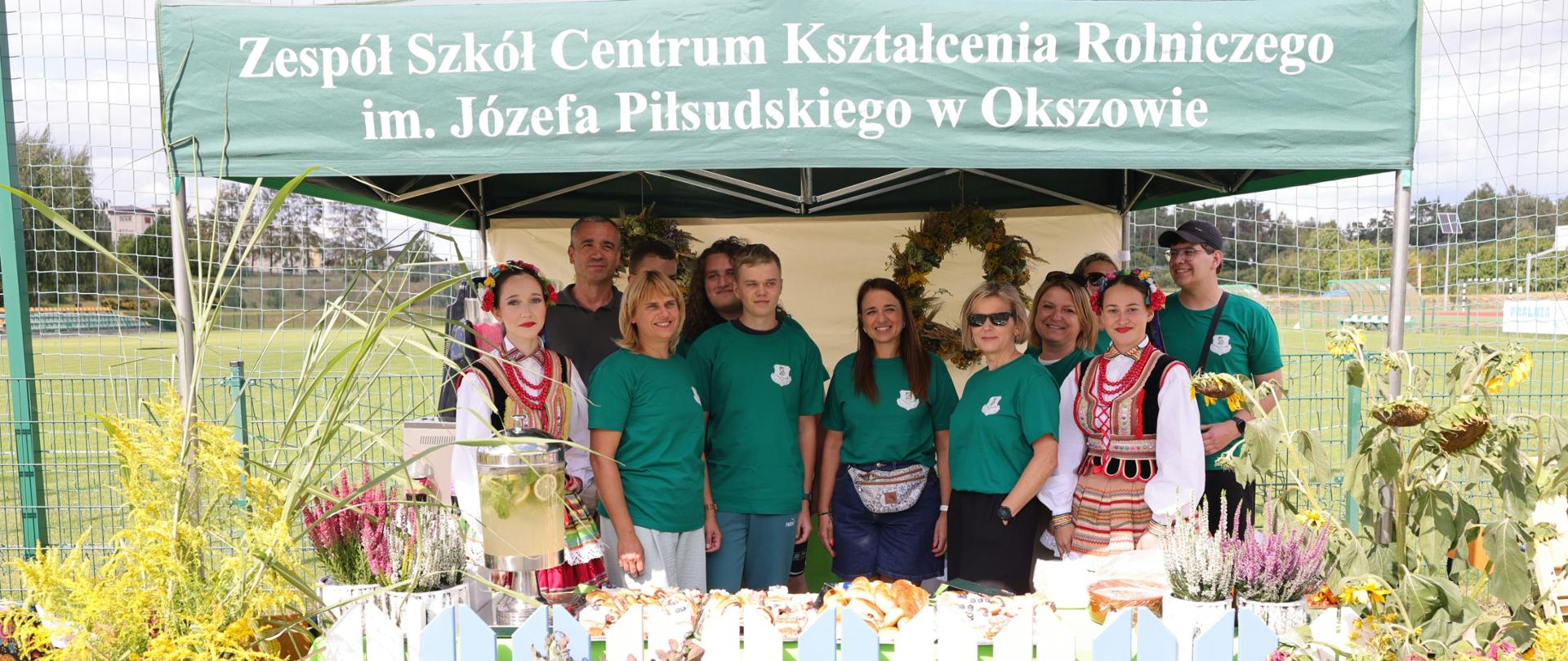 Grupa uczestników wydarzenia stoi przy stoisku ZSCKR w Okszowie udekorowanym kwiatami i roślinami.