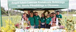 Grupa uczestników wydarzenia stoi przy stoisku ZSCKR w Okszowie udekorowanym kwiatami i roślinami.