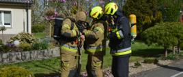 Ćwiczenia w DPS. Trzech strażaków na tle parkowej zieleni przygotowuje się do wejścia do budynku, w którym ma miejsce pożar. Zakładają sprzęt ochrony układu oddechowego. Dwóch z nich ma hełmy koloru żółtego z czerwonymi paskami, trzeci na głowie ma kominiarkę. Obok strażaków na chodniku leży rozwinięty wąż.