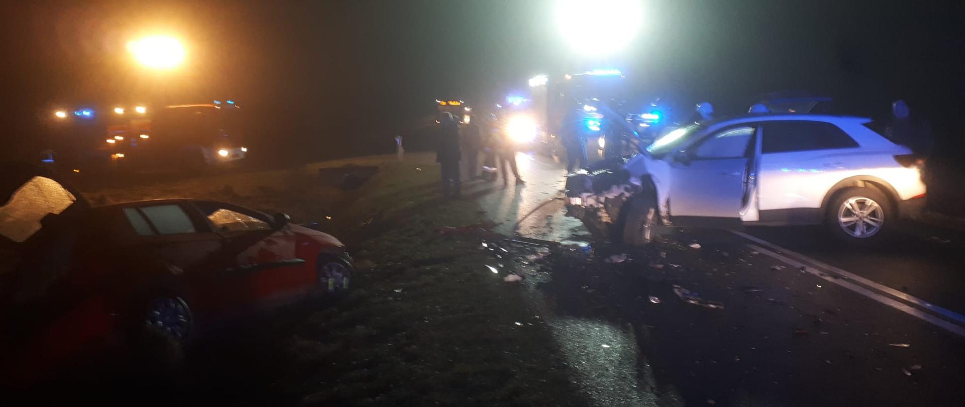 Zdjęcie przedstawia dwa z pośród trzech samochody, które brały udział w wypadku komunikacyjnym. W głębi stoją ratownicy i widać pojazdy służb ratowniczych.