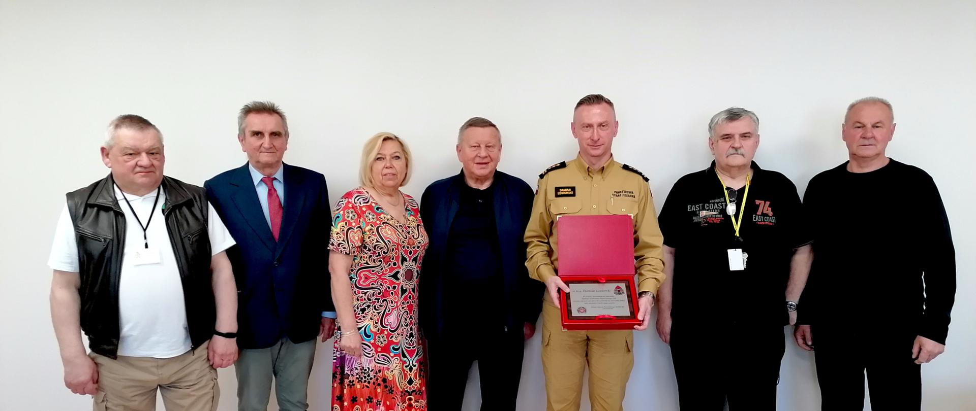 Na zdjęciu widzimy siedem osób stojących na tle białej ściany, pozujących do wspólnej fotografii grupowej. W centrum znajduje się śląski komendant wojewódzki starszy brygadier Damian Legierski w mundurze koloru piaskowego (beżowego), trzymający otwarte czerwone pudełko z odznaką i tabliczką pamiątkową. Pozostałe osoby to kobieta i sześciu mężczyzn członkowie Zarządu emerytów i rencistów, ubrani w różnorodne stroje – garnituru i sukienki, ubrania codzienne. Wszyscy stoją blisko siebie, uśmiechając się lekko.