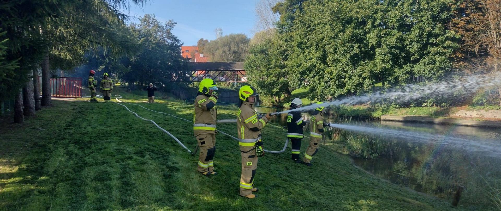 Strażacy ćwiczą podawanie wody nad brzegiem rzeki. Strażacy ubrani w mundury bojowe i żółte hełmy. Leją wodę z prądownic. Słonecznie.