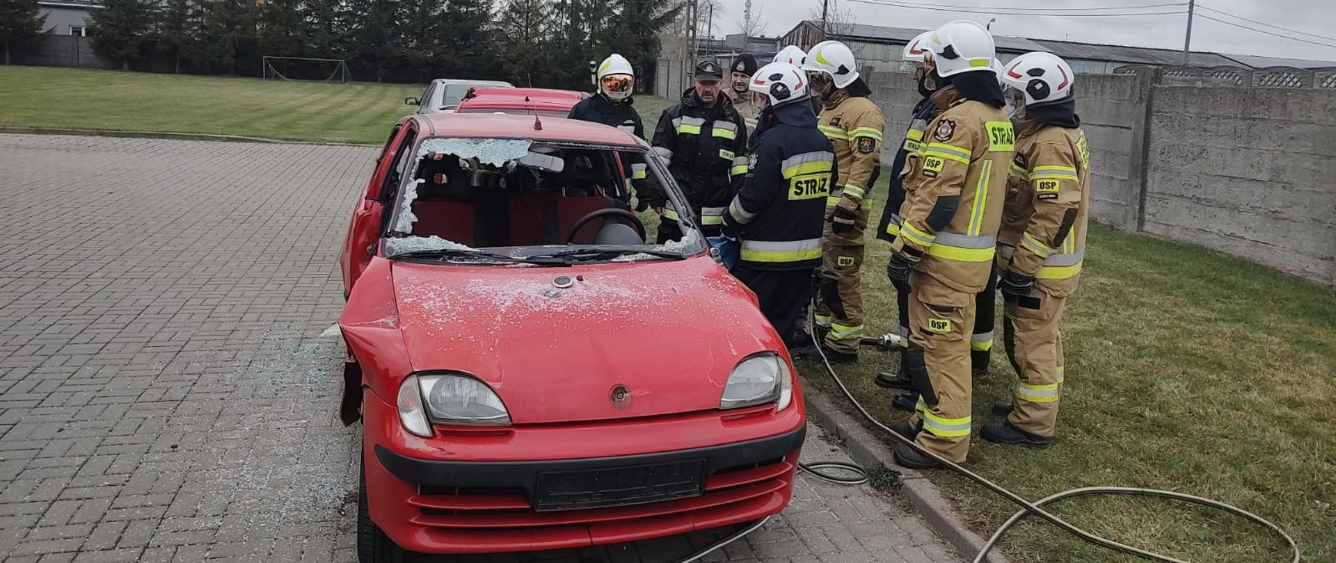 Strażacy w ubraniach specjalnych stoją przy czerwonym samochodzie osobowym, który będą cięli przy użyciu narzędzi hydraulicznych.