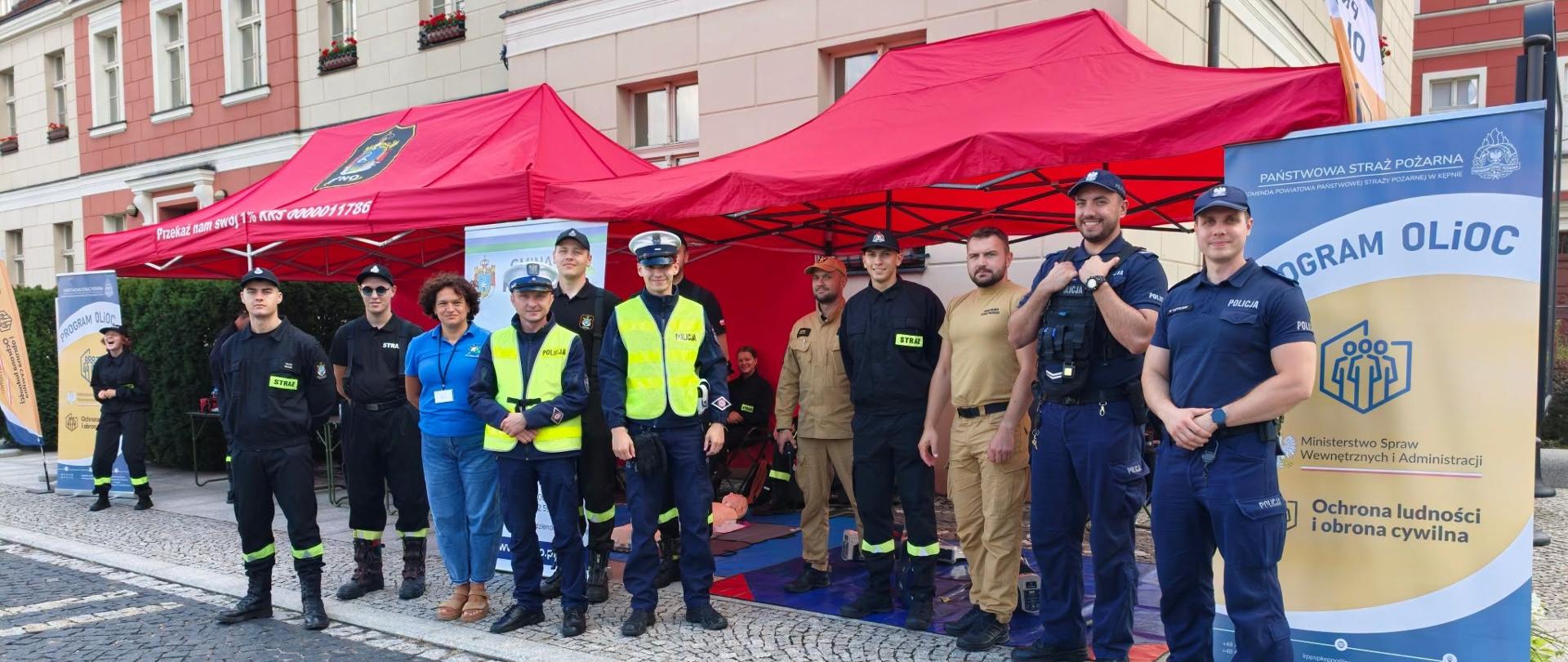 Zdjęcie przedstawia grupę około dziesięciu osób – funkcjonariuszy policji, strażaków oraz pracowników cywilnych – pozujących do pamiątkowej fotografii na brukowanym rynku miasta przed zabytkową kamienicą.
W tle stoją dwa czerwone namioty ogrodowe. Po prawej stronie ustawiono duży baner z napisem „PROGRAM OLiOC – Ochrona ludności i obrona cywilna” oraz logo Ministerstwa Spraw Wewnętrznych i Administracji i Państwowej Straży Pożarnej w Kępnie. Pod namiotami widoczny jest sprzęt do pierwszej pomocy, w tym fantom do resuscytacji leżący na macie.
Osoby na pierwszym planie stoją w jednym szeregu:
Po prawej stronie stoi dwóch postawnych policjantów w granatowych mundurach służbowych.
W środku i po lewej stronie znajdują się funkcjonariusze w odblaskowych kamizelkach policyjnych, strażacy w czarno-zielonych oraz piaskowych mundurach z naszywkami „STRAŻ”, a także kobieta w niebieskiej koszulce z identyfikatorem.
