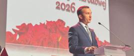 Wicewojewoda w granatowym garniturze i szarym krawacie przemawia przy mównicy. W tle wyświetlana jest grafika z napisem „Dzień Edukacji Narodowej 2026”, ozdobiona czerwonymi różami.