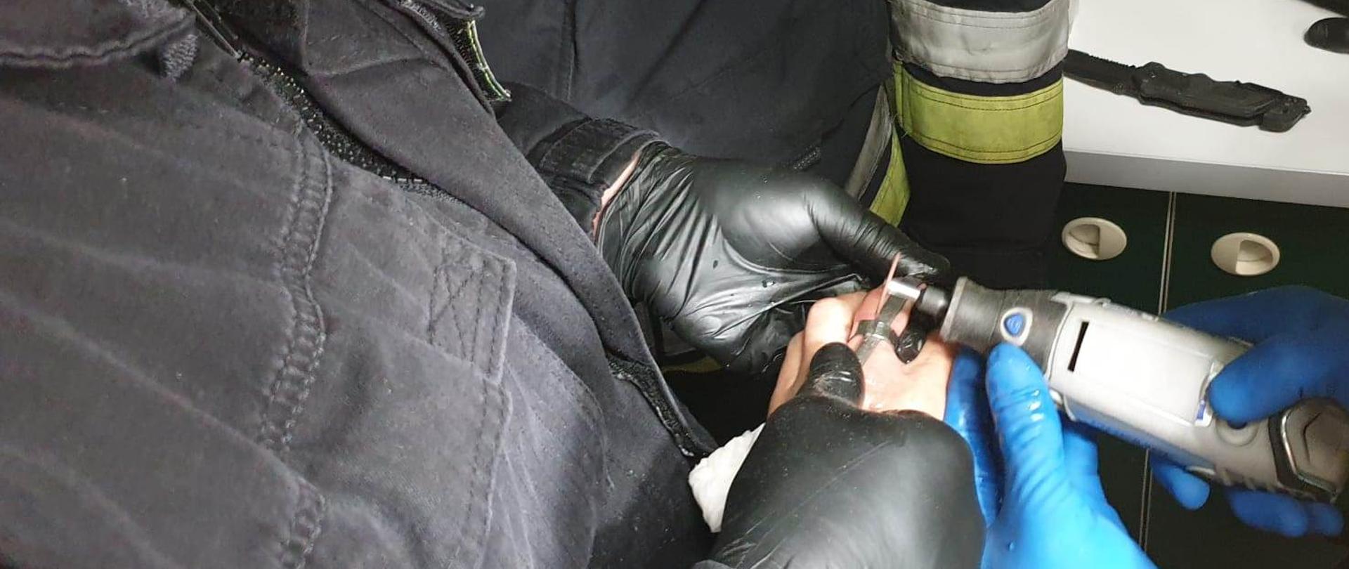 Zdjęcie obrazuje strażaków przecinających obrączkę na palcu kobiety, który spuchł i nie było możliwości jej ściągnięcia. Strażacy mają założone maseczki chirurgiczne oraz rękawiczki lateksowe. Obrączkę przecinają za pomocą szlifierki wielofunkcyjnej. W tle widać stół i przewody od urządzeń służących do pomiarów medycznych. 