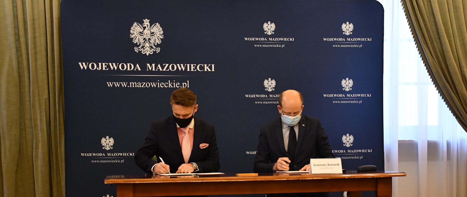 Zdjęcie przedstawia moment podpisania umowy. Za drewnianym stołem, na tle granatowej ścianki wizerunkowej Wojewody Mazowieckiego, siedzi dwóch mężczyzn ubranych w ciemne garnitury, koszule i krawaty.