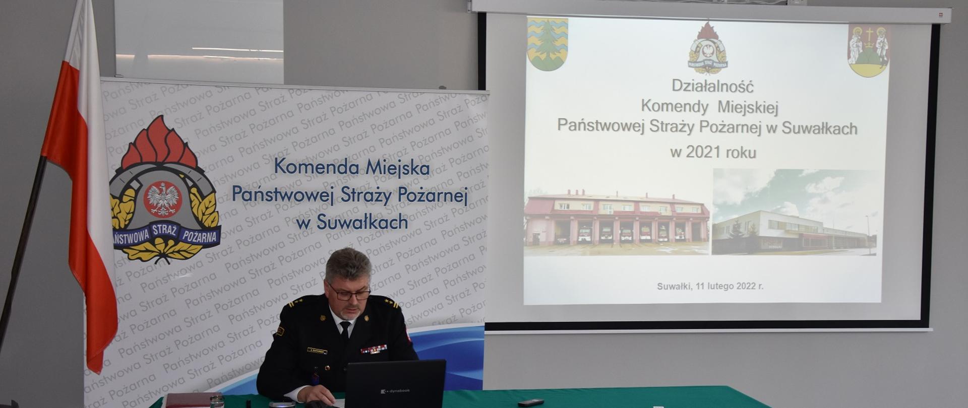 Zdjęcie obrazuje strażaka za biurkiem przy komputerze w tle flaga RP logo PSP z napisem Komenda Miejska Państwowej Straży Pożarnej w Suwałkach i planszą z prezentacją dotycząca działalności jednostek ochrony przeciwpożarowej powiatu suwalskiego w 2021 r. 